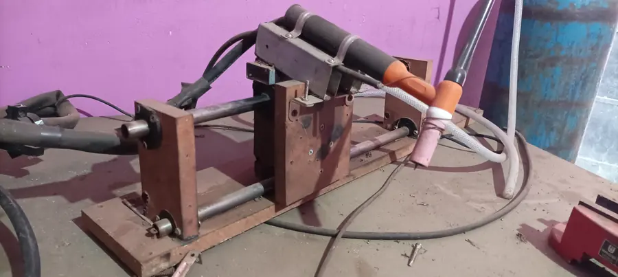 The welding arm on a workshop bench: a plywood and steel frame carrying the TIG torch with its ceramic cup, gas hose and cabling